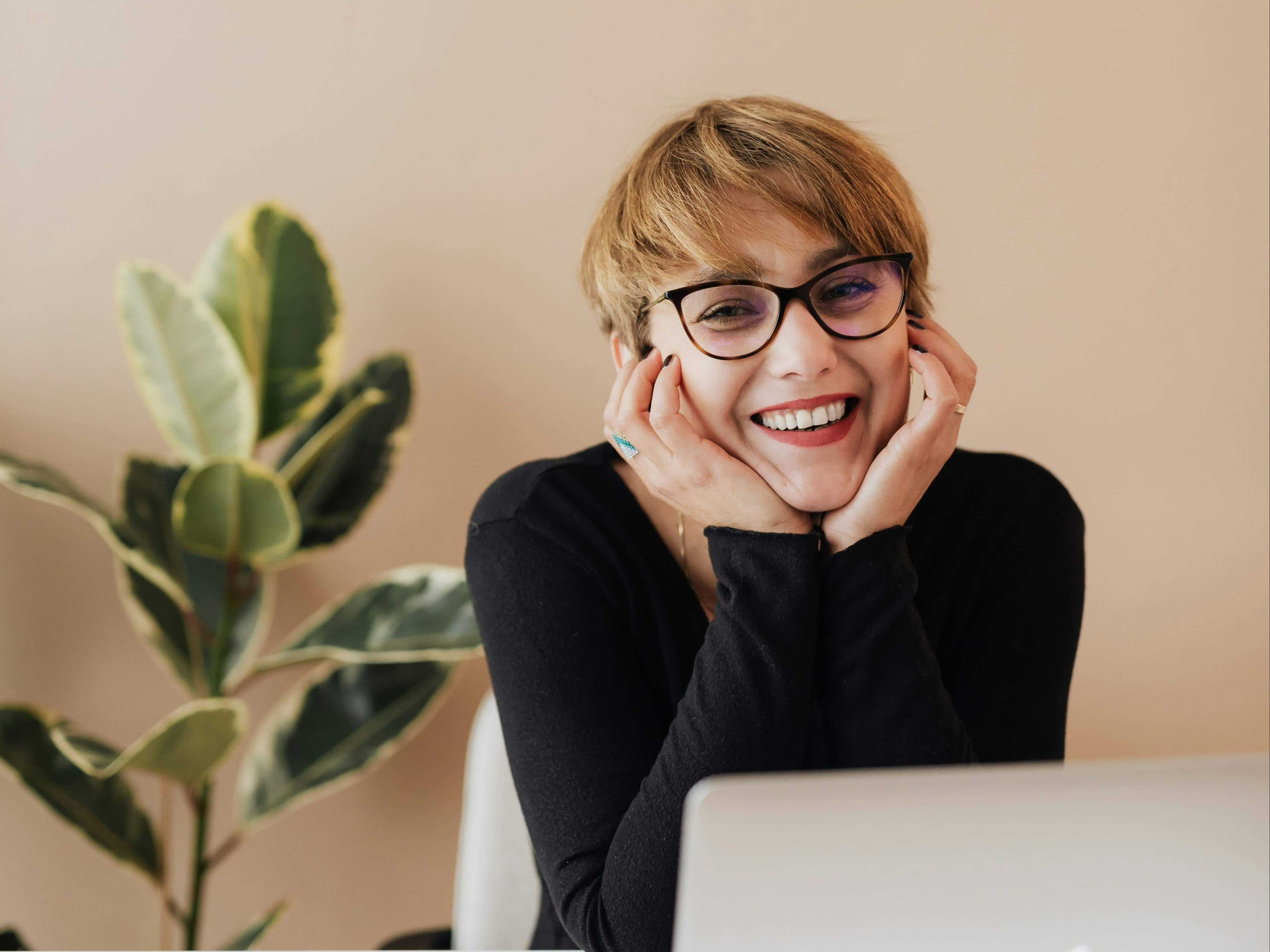A smiling woman with short hair, wearing glasses, resting her chin on her hands while sitting in front of a laptop with a plant in the background.