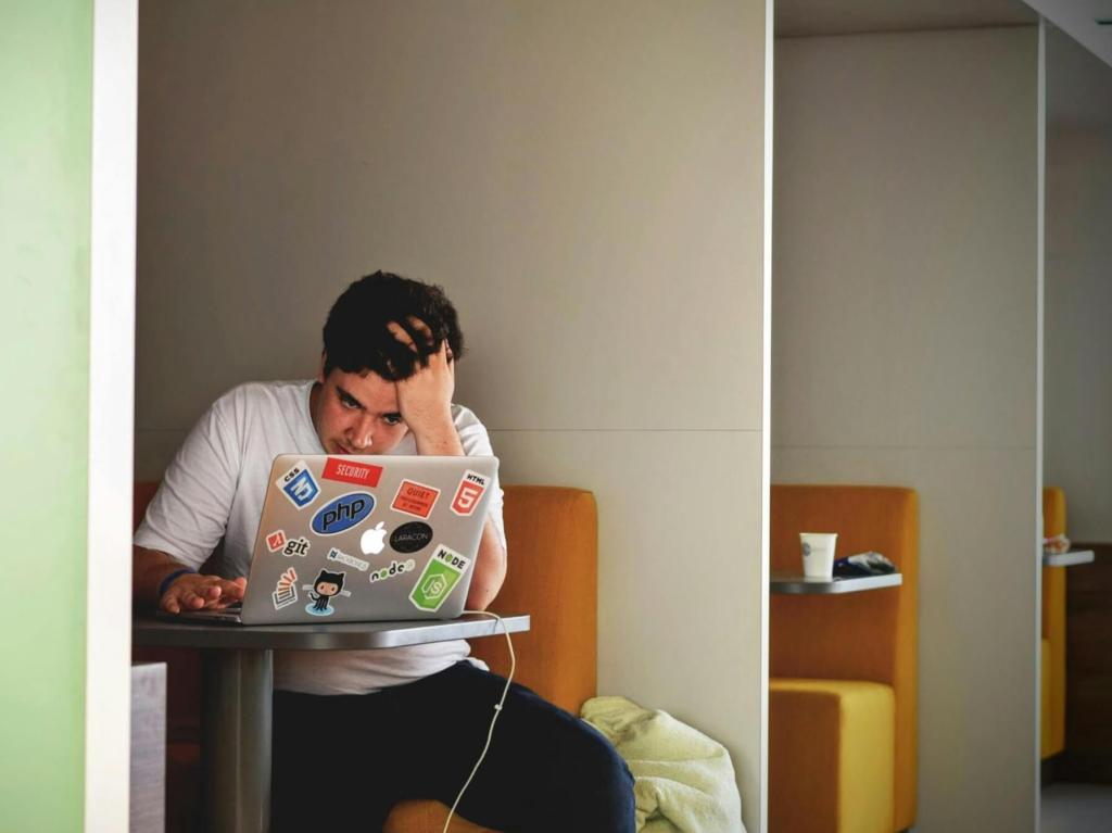A person sitting at a table, looking stressed while working on a laptop covered with various stickers.
